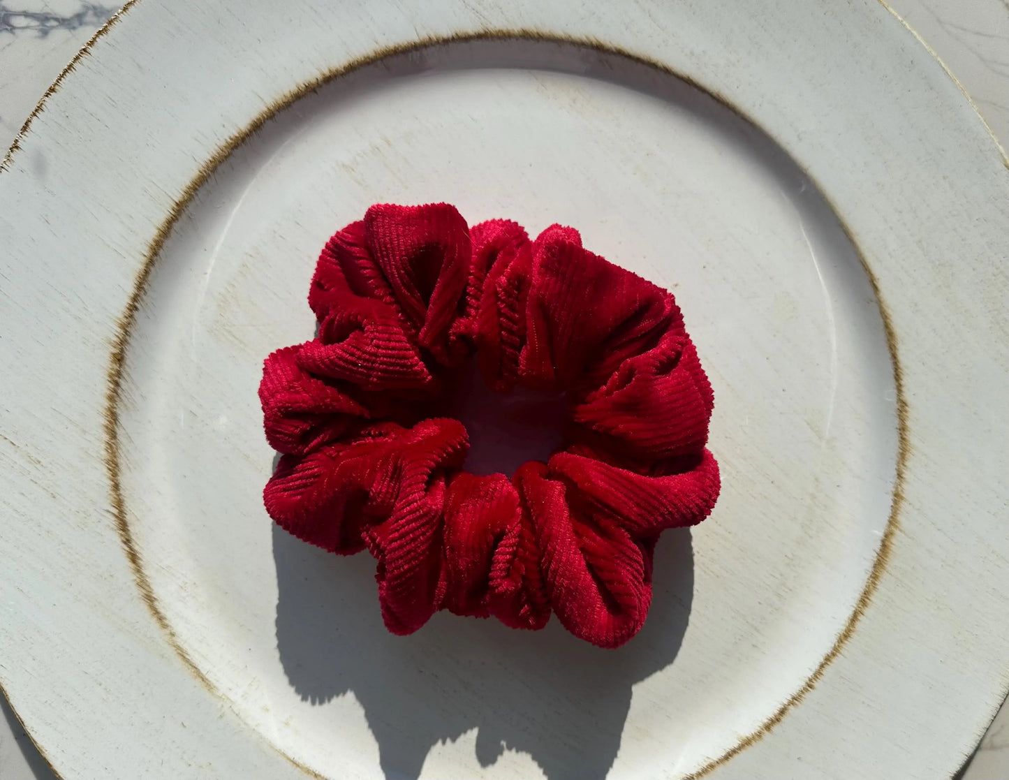 A deep red corduroy scrunchie placed on a white ceramic plate with a gold-toned rim, photographed from above in bright natural light.