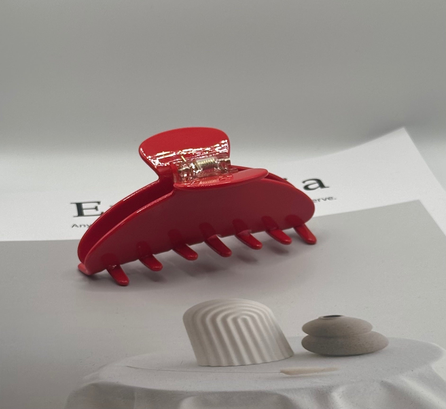 A glossy red hair claw clip with a gold-toned metal spring, displayed on a white surface with a white fabric-covered pedestal and two small decorative objects in the foreground.