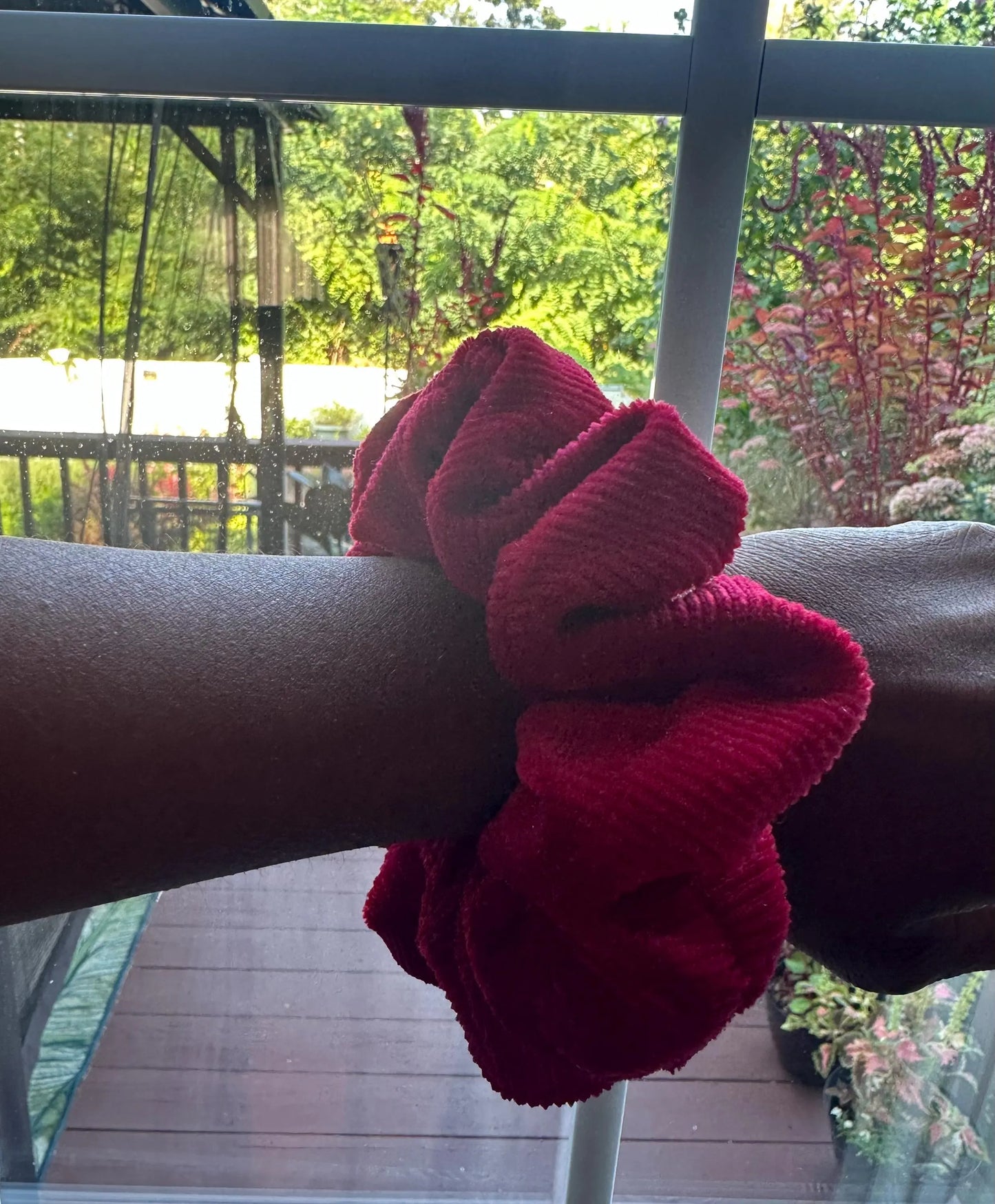 A dark red corduroy hair scrunchie worn on a person's wrist, with a balcony railing and green garden foliage visible in the background.