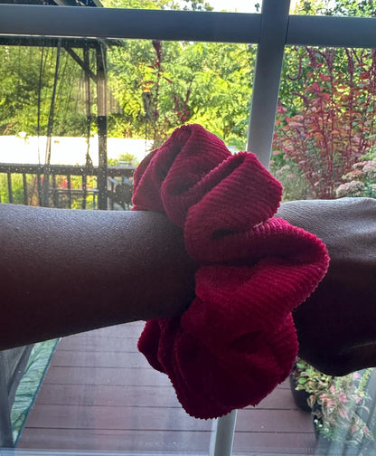A dark red corduroy hair scrunchie worn on a person's wrist, with a balcony railing and green garden foliage visible in the background.