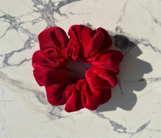 A deep red corduroy hair scrunchie resting on a white marble surface with gray veining, casting a soft shadow to the right.